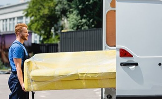 Side,View,Of,Loader,In,Uniform,Holding,Couch,In,Stretch Side view of loader in uniform holding couch in stretch wrap near truck with open doors on urban street